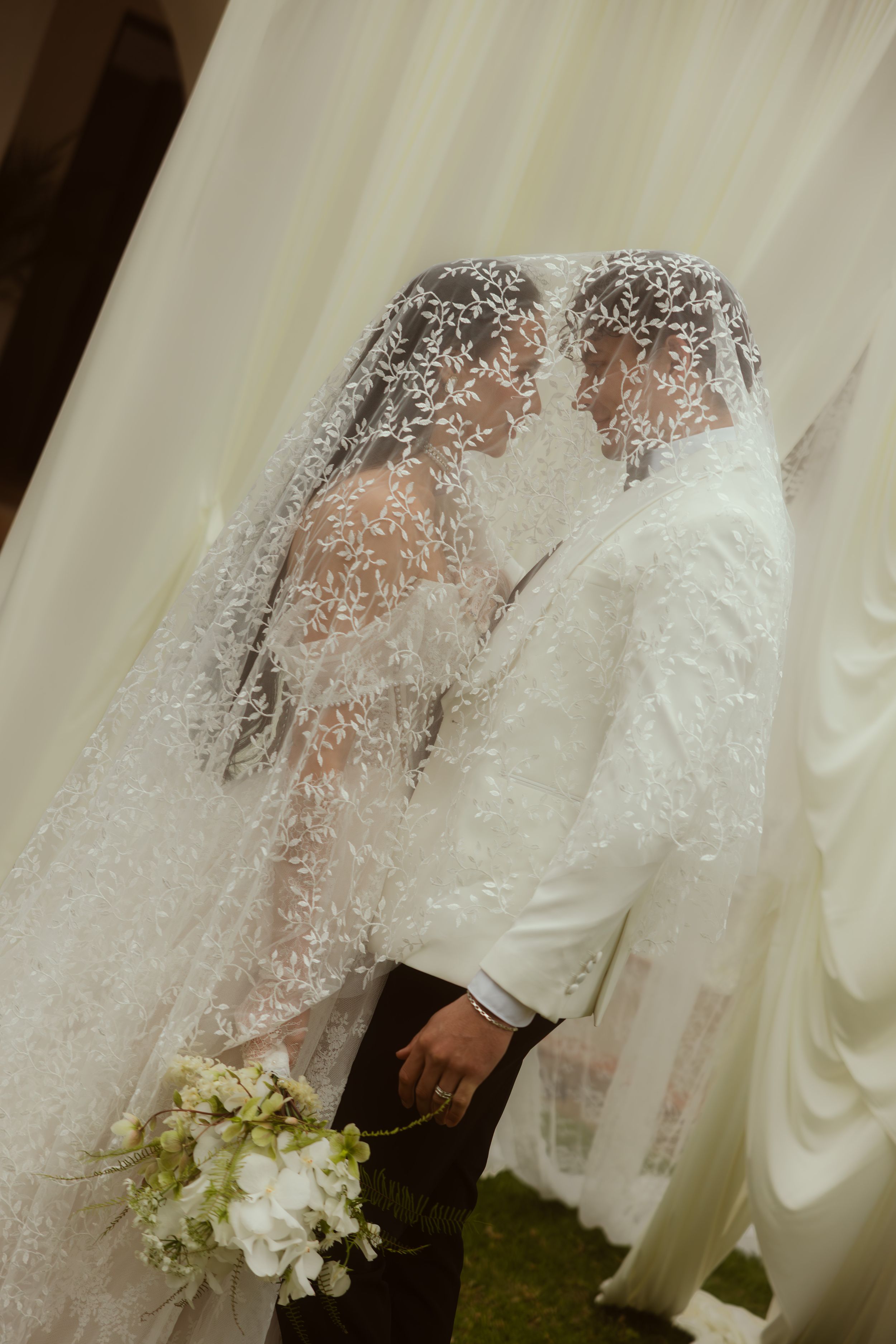 Bride and groom sharing a kiss under an intricate lace veil, holding white flowers at their wedding ceremony.