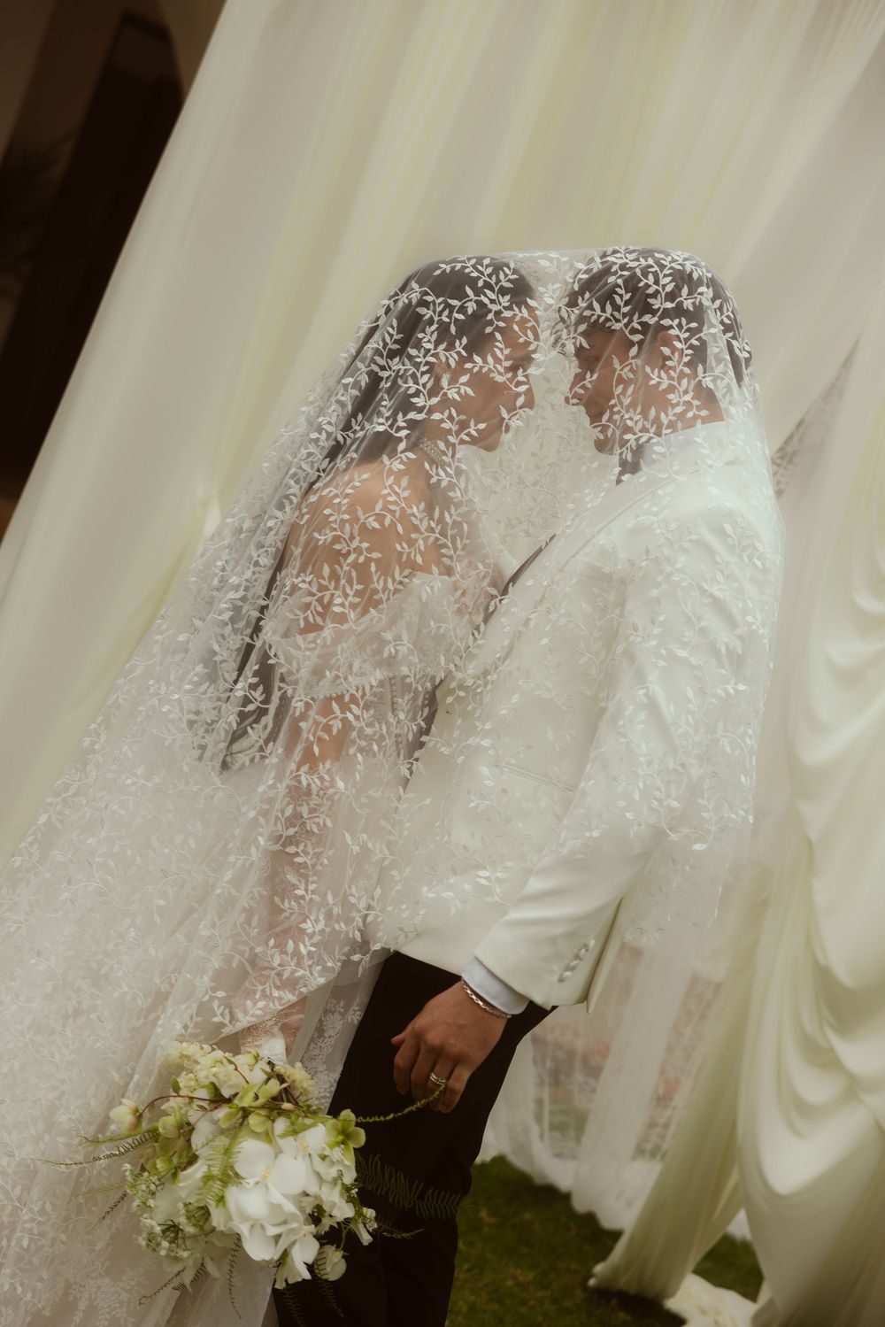Bride and groom sharing a kiss under an intricate lace veil, holding white flowers at their wedding ceremony.