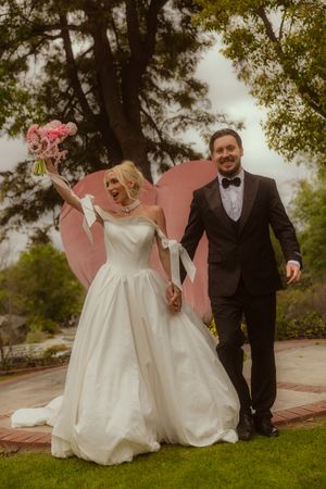 Joyful bride in white ball gown holding pink bouquet walks with groom in black tuxedo before heart backdrop outdoors.