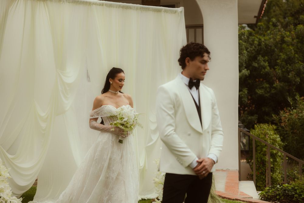 Bride in white off-shoulder gown and groom in white tuxedo stand before draped backdrop at outdoor wedding ceremony.