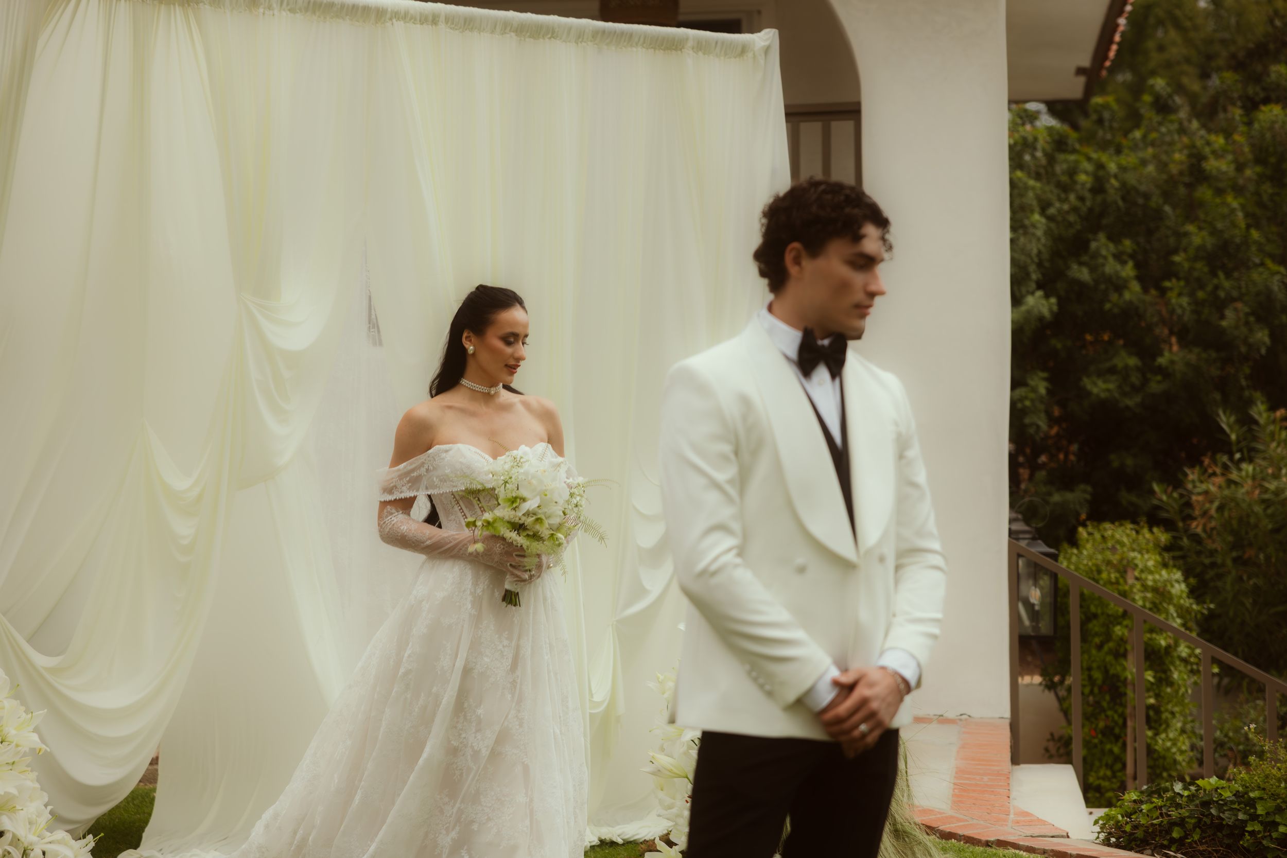 Bride in white off-shoulder gown and groom in white tuxedo stand before draped backdrop at outdoor wedding ceremony.