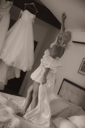 Bride in white off-shoulder gown jumping on bed with wedding dresses hanging in background, sepia tone.