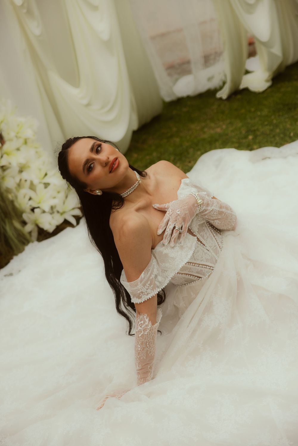 Elegant bride in lace off-shoulder wedding gown with gloves, sitting gracefully surrounded by white floral decor.