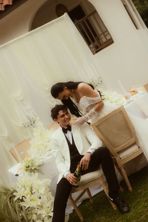 Bride and groom celebrate outdoors at elegant white-themed wedding reception, groom holding champagne bottle while seated.