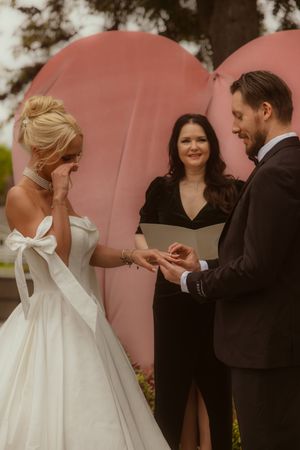 Emotional bride wiping tears as groom places ring on her finger during outdoor wedding ceremony with pink backdrop.