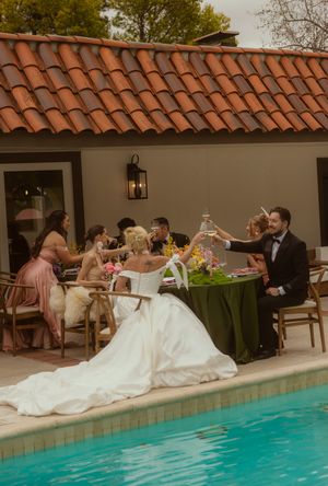 Bride and groom toast poolside with wedding party at elegant outdoor reception with terracotta roof.