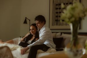 Couple sitting together on a bed in a dimly lit hotel room, man in white shirt embracing woman in black dress.