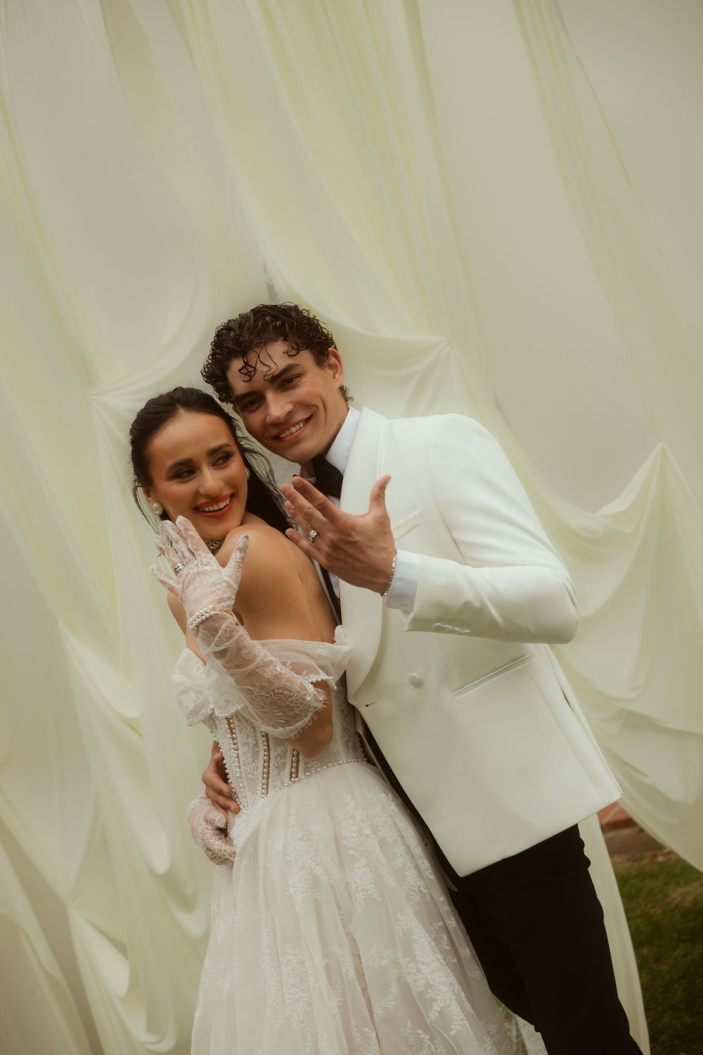 Bride and groom in white wedding attire pose together showing rings before flowing white curtains.