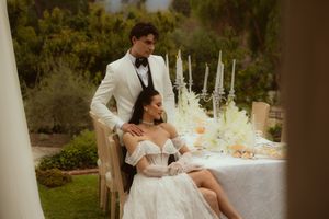 Elegant bride in off-shoulder gown seated at outdoor wedding table with groom in white tuxedo standing behind her.