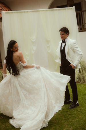 Bride in white lace ballgown twirls before groom in white tuxedo jacket against draped backdrop.