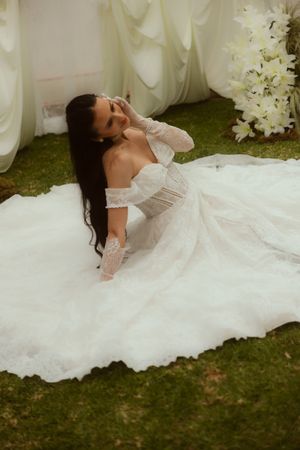 Bride in off-shoulder lace wedding gown with long train sits gracefully on green grass surrounded by white florals.