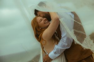 Joyful bride with red hair embraces groom under flowing white veil on their wedding day.