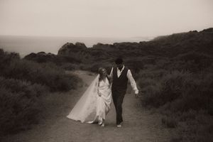 Bride and groom walk hand in hand along a coastal path in a romantic black and white wedding photo.