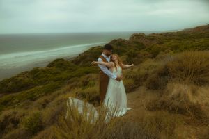 Couple embracing in wedding attire on a coastal cliff with lush greenery and ocean views on a cloudy day.