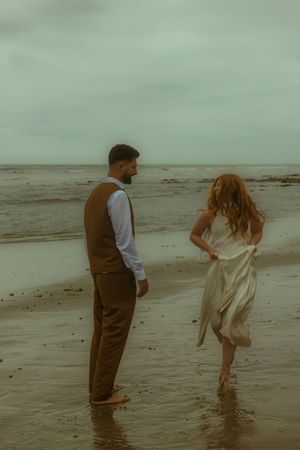 A couple stands barefoot on a sandy beach, the woman in a white dress and man in a brown vest, with moody overcast skies.
