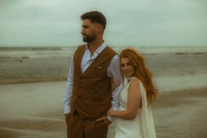 Couple posing on a beach, man in brown vest suit, woman in white dress with red hair, overcast sky.