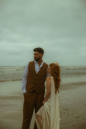A bride and groom stand on a sandy beach under a cloudy sky, she in a white gown, he in a brown vest.