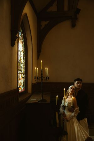 Bride and groom stand by candlelight in a gothic chapel with stained glass windows during a romantic wedding ceremony.