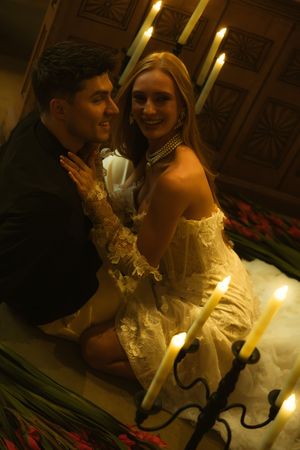 Romantic couple sitting among candles in warm golden light, woman in white lace dress smiling, intimate candlelit setting.