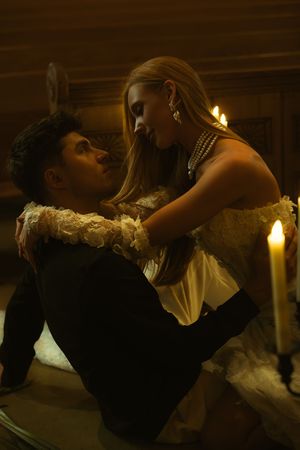 A couple shares a romantic candlelit moment, woman in white dress with pearls gazing down at man in dark suit.