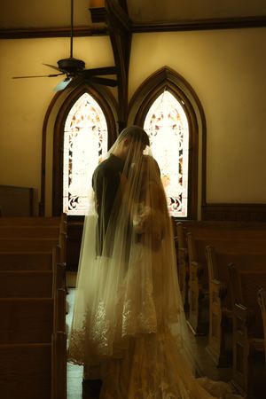 Bride in lace wedding gown and veil stands before stained glass windows in a wooden church aisle.