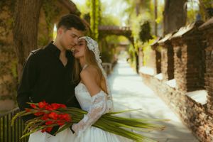 Bride and groom share a romantic moment on a garden path, she holds red tropical flowers in a lace wedding gown.
