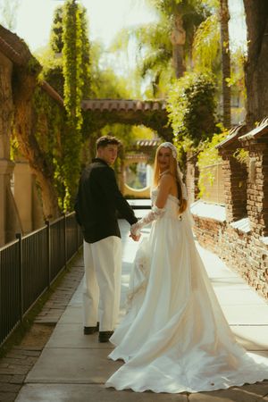 A bride in a flowing white gown and groom in black suit hold hands on a sunlit garden pathway.