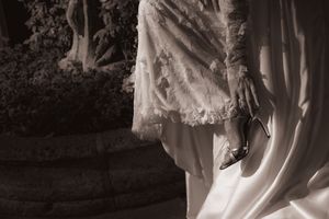 Sepia-toned bride in lace gown holding heels near stone fountain, elegant wedding photography.
