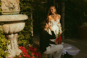 Man kneeling before woman in white dress during romantic garden proposal near stone fountain with red flowers.