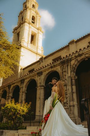 Bride and groom embrace in front of a Spanish colonial church with bell tower and arched colonnade on a sunny day.