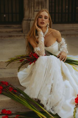 Bride in white lace gown with floral gloves sits gracefully among red gladiolus flowers outdoors.