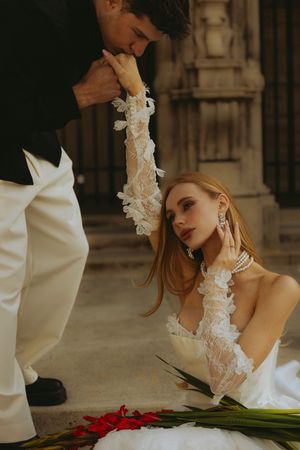 Groom kissing bride's lace gloved hand as she sits gracefully in a white wedding gown with pearl jewelry.