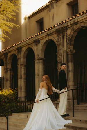 Bride in off-shoulder white gown and groom in black suit on steps of elegant arched stone building at golden hour.