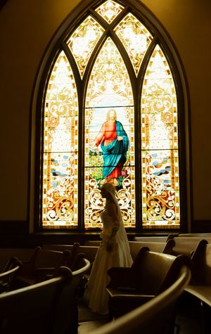 Bride in white gown standing before a stunning stained glass window depicting Virgin Mary in a historic church.