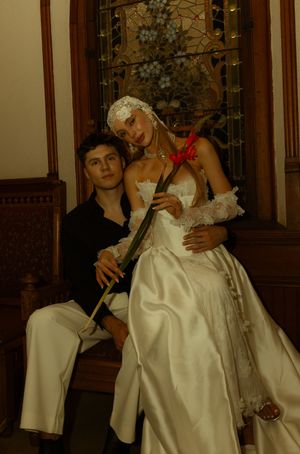 A couple in formal attire poses in a chapel, bride in white gown with veil holding a red flower.