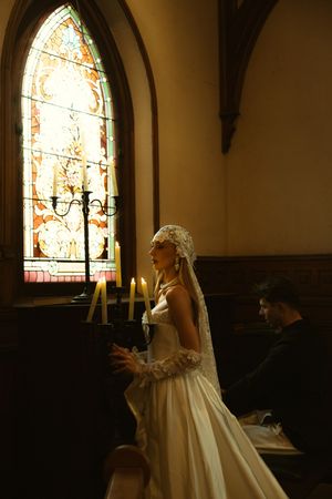 Bride in white gown and lace veil stands by candlelight near stained glass window in a church.