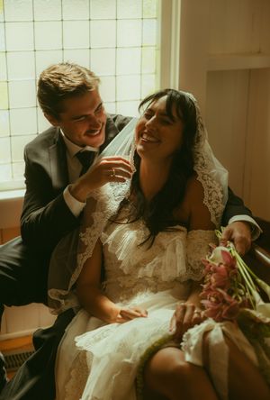 Happy bride in lace veil holding pink flowers laughs with smiling groom in dark suit during wedding ceremony.