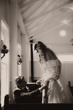Black and white photo of a groom kneeling before his bride in a wedding gown and veil inside a chapel.