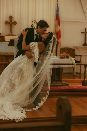Bride and groom share a romantic dip kiss at the church altar, bride wearing a lace wedding gown with cathedral veil.
