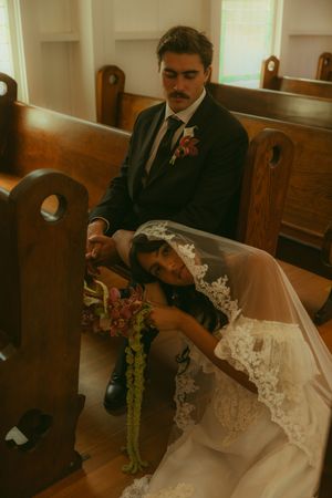 Bride in lace wedding dress rests head on groom's lap while sitting in church pews, holding floral bouquet.