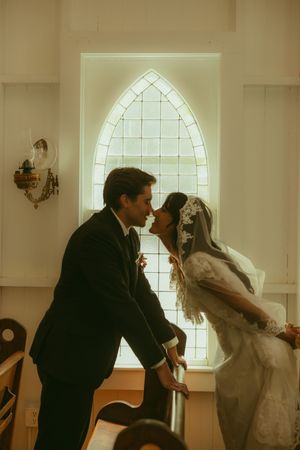 A bride and groom share a romantic kiss in a sunlit chapel with a Gothic arched stained glass window behind them.