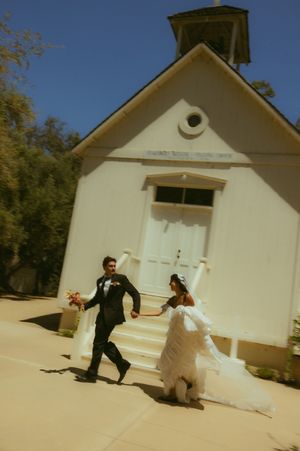 Newlywed couple holding hands walking outside a white chapel on their wedding day under a clear blue sky.