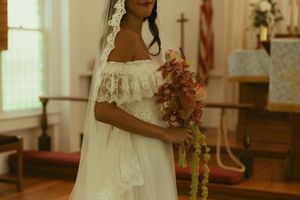 Bride in white lace off-shoulder gown holding floral bouquet inside a church.