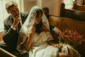 Bride in lace veil and groom in gray suit sit together in a church pew during their wedding ceremony.
