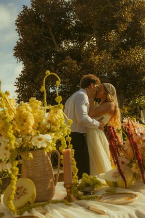 A couple shares a romantic kiss at an outdoor wedding reception table adorned with yellow flowers and candles.