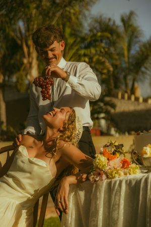 Groom feeds bride grapes at outdoor wedding reception table with tropical palm trees and floral arrangements.