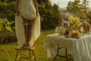 Bride in white dress standing on wooden stool beside elegant outdoor wedding table with floral arrangements and cake.
