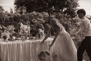 Black and white wedding photo of bride in white gown leaning over table with floral arrangements outdoors.
