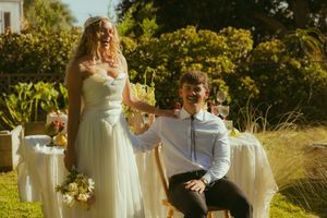 Bride in white strapless gown holding bouquet stands beside groom in suit toasting at outdoor wedding reception.