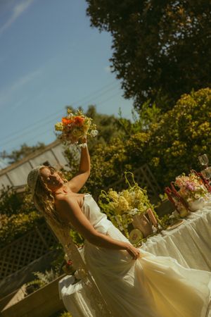 Bride in white gown raises colorful bouquet outdoors at sunny garden wedding reception table with floral arrangements.